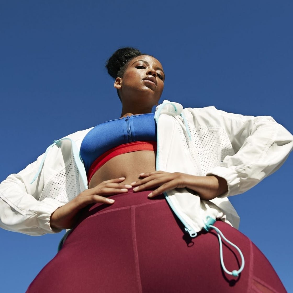 A woman wearing purple leggings, a blue sports bra and white jacket stood outside.